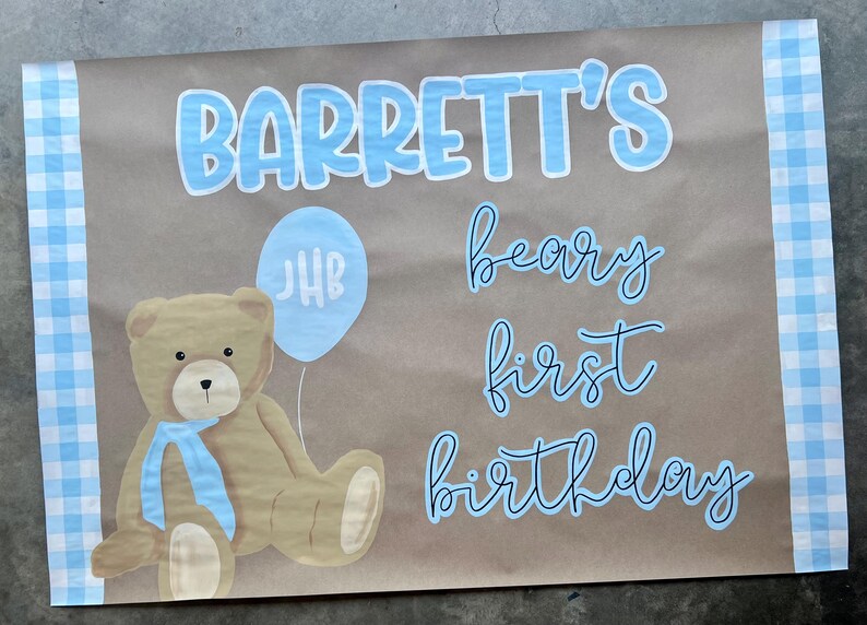 K&ouml;nnte beinhalten: Braunes Papierschild mit blauem Text: "Barrett's beary first birthday". Ein Teddyb&auml;r mit blauem Band und ein blauer Ballon mit den Initialen "JHB" sind ebenfalls abgebildet. Die R&auml;nder haben ein blau-wei&szlig;es Karomuster.