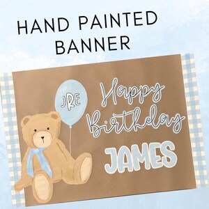 K&ouml;nnte beinhalten: Handgemaltes Geburtstagsbanner mit einem braunen Teddyb&auml;ren, einem hellblauen Ballon und den Worten "Happy Birthday JAMES". Das Banner hat einen braunen Hintergrund mit einem hellblau-wei&szlig;en Vichy-Rand.