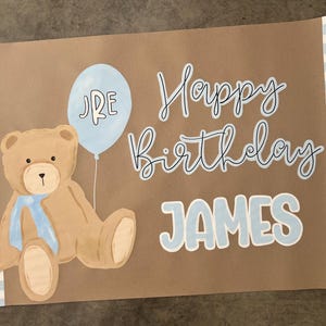 K&ouml;nnte beinhalten: Braune Papier-Geburtstagskarte mit einer Illustration eines hellblauen Ballons und Teddyb&auml;ren. Die Karte tr&auml;gt die Aufschrift "Happy Birthday JAMES" in blauer Schrift. Die Karte hat einen blau-wei&szlig; karierten Rand.