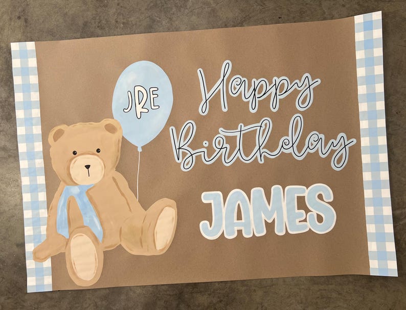 K&ouml;nnte beinhalten: Braune Papier-Geburtstagskarte mit einer Illustration eines hellblauen Ballons und Teddyb&auml;ren. Die Karte tr&auml;gt die Aufschrift "Happy Birthday JAMES" in blauer Schrift. Die Karte hat einen blau-wei&szlig; karierten Rand.