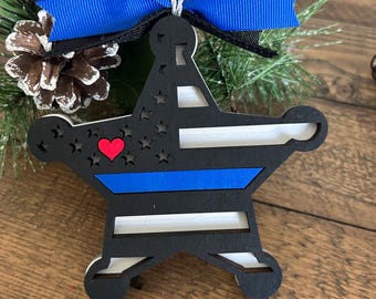 Könnte beinhalten: Ein schwarzer Holzstern-Ornament mit blauem Streifen und rotem Herz. Das Ornament hat ein weißes und schwarzes Flaggenmuster. Es ist mit einem blauen Band und Tannennadeln verziert.