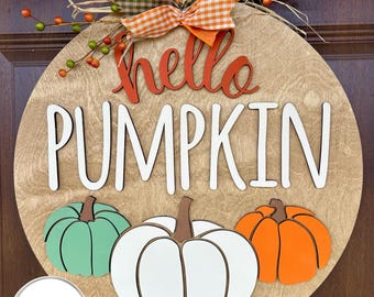 May include: A round wooden door sign featuring the text hello PUMPKIN in orange and white. Below the text are three wooden pumpkins in light green, white, and orange. The sign is decorated with a plaid bow and artificial berry sprigs.