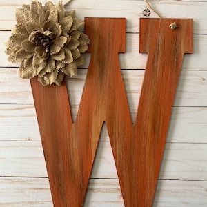 Könnte beinhalten: Ein Holzbuchstabe W, der in Orange- und Brauntönen bemalt ist und eine Burlap-Blume oben befestigt hat. Der Buchstabe hängt an einer Schnur.