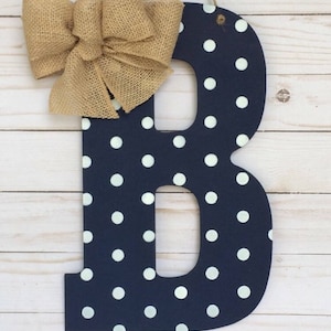 Puede incluir: Una letra B de madera azul marino con lunares blancos y un lazo de arpillera. La letra está colgada de una cuerda.