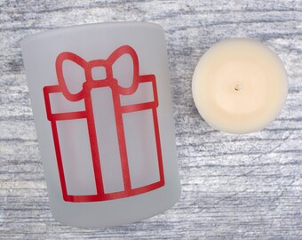 May include: A frosted glass votive candle holder with a red gift box design and a cream-colored candle. The candle holder is cylindrical and the gift box design is outlined in red. The candle is round and has a wick.