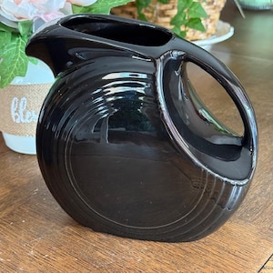 May include: A glossy black ceramic pitcher with a rounded body and a large, open handle. The pitcher has a curved spout and a ridged design around the base. The pitcher is likely used for serving beverages.