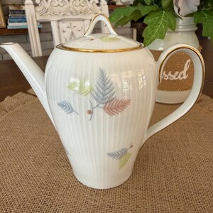 May include: White porcelain teapot with a lid, featuring a gold rim and handle. The teapot is decorated with a leaf pattern in pastel colors. The teapot has a long spout and a curved handle.