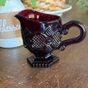 Pode incluir: Um cremeira de vidro vermelho rubi com uma alça decorativa e detalhes em relevo ornamentados. A cremeira tem uma base quadrada e um bico. O design apresenta padrões geométricos e uma superfície texturizada, adicionando charme vintage.