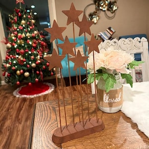 May include: A decorative metal star ornament featuring seven stars of different sizes. The stars are fixed to thin rods and set on a rectangular base. A Christmas tree and a vase of flowers are in the background.