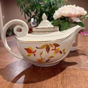 Puede incluir: Tetera de cerámica color crema con tapa y asa curva. La tetera presenta un diseño floral en tonos amarillos, naranjas y marrones, con ribete dorado. La tetera está sobre una superficie de madera.