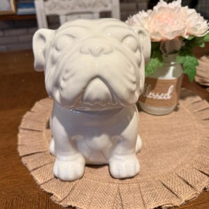Op de afbeelding: Een witte keramische bulldogfiguur met een glanzende afwerking. De bulldog zit rechtop met zijn voorpoten naar voren. Het beeldje staat op een beige placemat. Een klein glazen potje met het woord "blessed" staat op de achtergrond.
