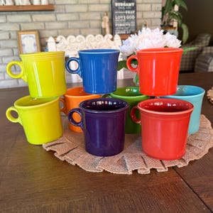 May include: A collection of nine ceramic mugs in various solid colors including lime green, cobalt blue, bright orange, deep purple, grass green, turquoise, and red. Each mug features a smooth finish, a rounded handle, and a consistent cylindrical shape.