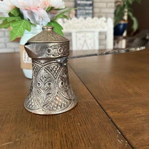 Può includere: Una caffettiera argentata ornata con un lungo manico. La caffettiera presenta un motivo floreale decorativo e un coperchio. È appoggiata su un tavolo di legno. Un vaso di fiori è sullo sfondo. La caffettiera è alta circa 15 cm.