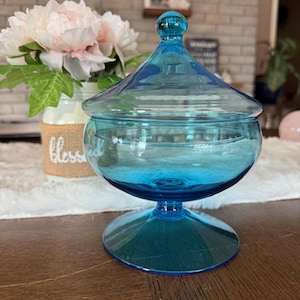 Könnte beinhalten: Eine klare, türkisfarbene Bonbonniere aus Glas mit Deckel. Die Schale hat eine runde Schalenform, einen Sockel und einen abgerundeten Knopf auf dem Deckel. Die Schale steht auf einer Holzoberfläche.