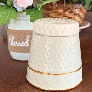 Op de afbeelding: Crèmekleurige keramische pot met een textuur en een gouden band. De pot heeft een deksel met een kuiltjespatroon. Een witte pot met het woord "blessed" staat op de achtergrond. De pot is een decoratief item.