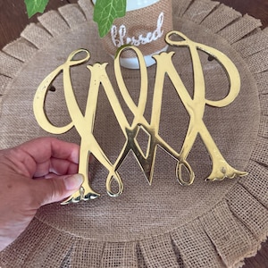 May include: Gold-coloured metal monogram featuring the letters JMP intertwined. The monogram is displayed on a burlap placemat with a ruffled edge. A white ceramic pot with the word "Blessed" is in the background.