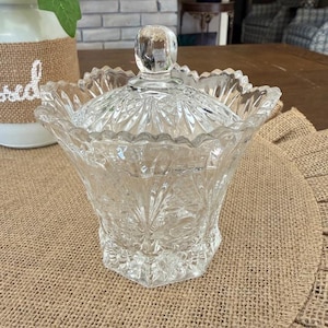 Peut inclure: Un bonbonnière en verre taillé transparent avec un couvercle. Le plat a un bord festonné et un bouton à facettes sur le couvercle. Le verre est orné d'un motif floral. Le plat est posé sur un dessous de plat en toile de jute.