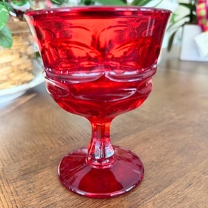 Puede incluir: Una copa de vidrio rojo rubí con un detallado diseño floral. La copa tiene un cuenco redondeado, un tallo corto y una base circular. El vidrio refleja la luz, resaltando el intrincado diseño. Probablemente se utiliza para servir bebidas.
