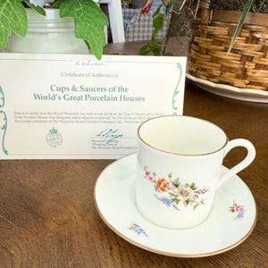 Puede incluir: Un juego de taza y platillo de porcelana blanca con un diseño floral y ribete dorado. El juego va acompañado de un certificado de autenticidad. La taza y el platillo están sobre una superficie de madera.