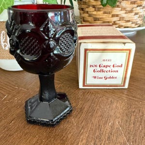 May include: A ruby red wine goblet from the 1876 Cape Cod Collection. The glass features an ornate design with a textured pattern and a hexagonal base. The original box is also visible.