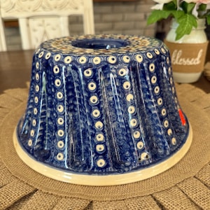 Puede incluir: Un molde para pastel bundt de cerámica azul y blanca con un patrón repetitivo de pequeños círculos y puntos. El exterior tiene estrías verticales y un acabado brillante, con un orificio central y un borde base ancho y liso de color crema.