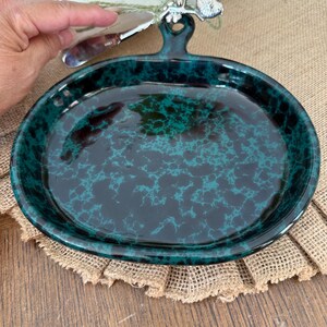 Può includere: Un piatto rotondo in ceramica con una smaltatura lucida e maculata nei toni del verde acqua scuro e del nero. Il piatto presenta una piccola impugnatura rialzata sulla parte superiore con un foro per appenderlo. La superficie è liscia.