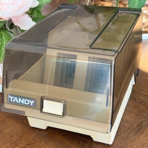 Könnte beinhalten: Ein Vintage Tandy CD-Aufbewahrungskoffer. Der Koffer besteht aus beigem und klarem Kunststoff mit einem klappbaren, getönten Deckel. Das Wort "TANDY" ist auf der Vorderseite aufgedruckt. Der Koffer hat einen weißen Sockel und einen rechteckigen Knopf.