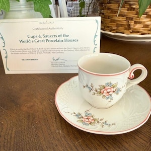 May include: A white porcelain teacup and saucer set with a floral design in pink and blue. The set includes a certificate of authenticity. The cup and saucer have a delicate, vintage aesthetic.