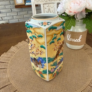 May include: A decorative, hexagonal vase with a detailed, hand-painted landscape scene. The vase features a yellow background with images of birds, flowers, and water. The top rim has a gold trim. The vase is sitting on a burlap placemat.