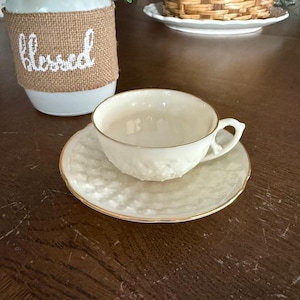 Könnte beinhalten: Cremefarbenes Teetassen- und Untertassen-Set mit strukturierter Oberfläche und goldenem Rand. Die Teetasse hat einen zarten Henkel. Die Untertasse ist rund. Das Set steht auf einem dunkelbraunen Holztisch. Ein weißes Glas mit dem Wort "Blessed" ist im Hintergrund.