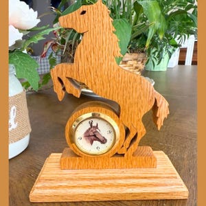 May include: A wooden clock featuring a rearing horse silhouette. The clock face, with a horse image, is set within a gold-colored frame. The clock is mounted on a rectangular wooden base, showcasing a natural wood grain.