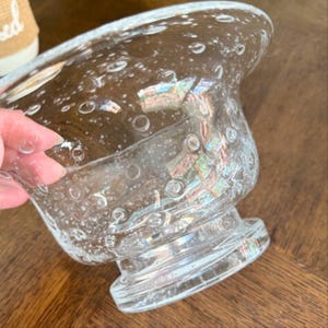 Puede incluir: Un cuenco de vidrio transparente con base de pedestal. El cuenco tiene una forma ancha y redondeada con una superficie texturizada y numerosas burbujas de aire. El vidrio refleja la luz, creando colores iridiscentes. El cuenco es probablemente decorativo.