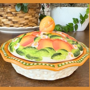 Puede incluir: Un plato de cerámica con temática de melocotón con tapa. La tapa está decorada con rodajas de melocotón, hojas verdes y pequeñas flores blancas. El plato tiene un borde naranja y un diseño de tejido de cesta en la base.