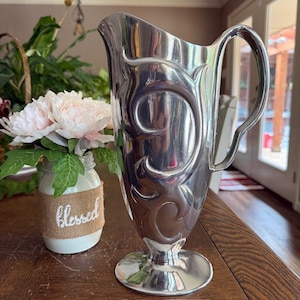 May include: A silver-colored pitcher with an ornate, swirling design. The pitcher has a handle and a pedestal base. Beside it is a small jar with the word "blessed" on it, holding pink flowers.