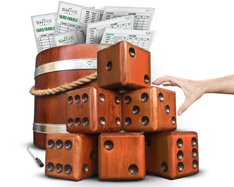 May include: A set of six large wooden dice in a brown wood finish. The dice are stacked in a pyramid shape with a hand reaching for the top die. The dice are for a yard game and come with scorecards.