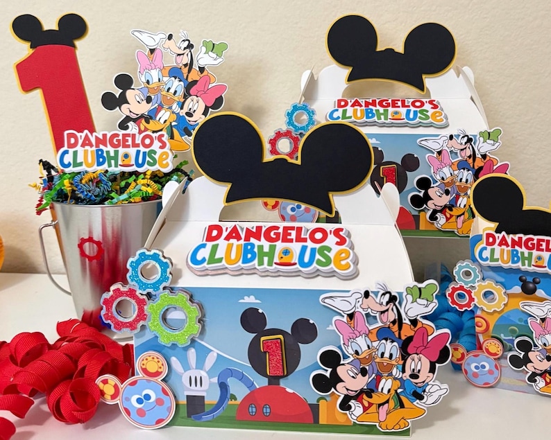 May include: Party decorations featuring Mickey Mouse and friends. Includes boxes with Mickey Mouse ears, the text "D'Angelo's Clubhouse", and character cutouts. A red number one and a metal bucket with colourful filler are also present.