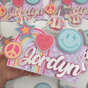 May include: A pink and white party favor box with a rainbow design and the name Jordyn in silver glitter letters. The box has a peace sign, a star, a smiley face, and a heart with the words "Girl Power" on it.