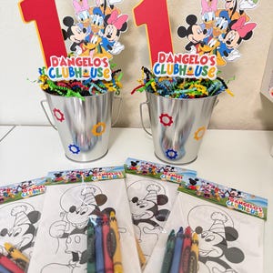 May include: Two silver metal buckets with colourful confetti and Mickey Mouse-themed decorations. Each bucket has a red number one and a sign that reads "D'Angelo's Clubhouse." Colouring pages with crayons are in front of the buckets.