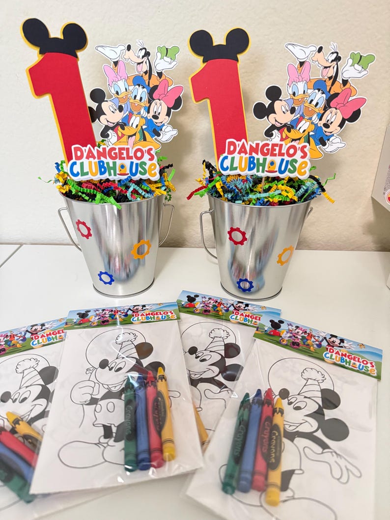 May include: Two silver metal buckets with colourful confetti and Mickey Mouse-themed decorations. Each bucket has a red number one and a sign that reads "D'Angelo's Clubhouse." Colouring pages with crayons are in front of the buckets.