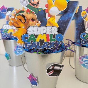 May include: A silver metal bucket centrepiece featuring a cutout of Luigi and a yellow dinosaur, with a sign reading Super Camilo Galaxy. The bucket is decorated with stickers of a Bullet Bill and a star, filled with blue and purple paper shreds.