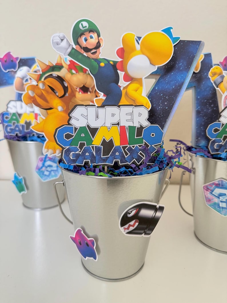 May include: A silver metal bucket centrepiece featuring a cutout of Luigi and a yellow dinosaur, with a sign reading Super Camilo Galaxy. The bucket is decorated with stickers of a Bullet Bill and a star, filled with blue and purple paper shreds.