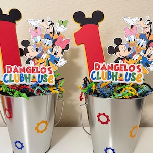 May include: Two silver metal buckets decorated with colourful gear shapes and filled with shredded paper. Each bucket has a red number one and a Mickey Mouse Clubhouse character cutout. The text "D'ANGELO'S CLUBHOUSE" is displayed on each bucket.