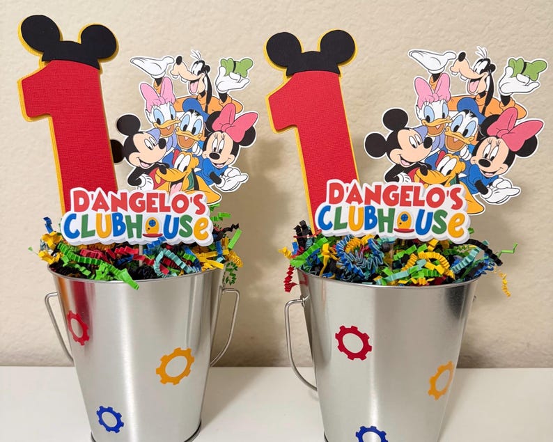May include: Two silver metal buckets decorated with colourful gear shapes and filled with shredded paper. Each bucket has a red number one and a Mickey Mouse Clubhouse character cutout. The text "D'ANGELO'S CLUBHOUSE" is displayed on each bucket.