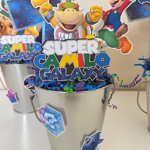 May include: A silver metal bucket centrepiece featuring cutouts of Bowser Jr., Mario, and a yellow star. The central sign reads Super Camilo Galaxy in a stylised font. The bucket is filled with purple and green paper shreds and decorated with stickers.