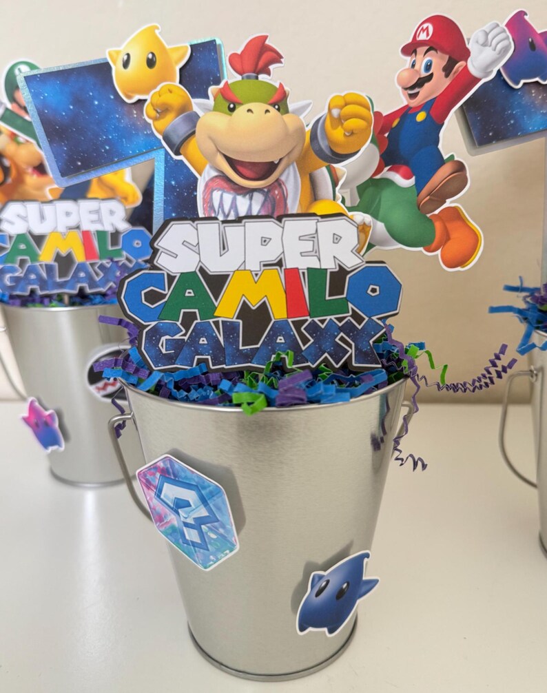 May include: A silver metal bucket centrepiece featuring cutouts of Bowser Jr., Mario, and a yellow star. The central sign reads Super Camilo Galaxy in a stylised font. The bucket is filled with purple and green paper shreds and decorated with stickers.