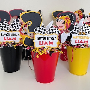May include: Three Mickey Mouse themed centerpieces for a birthday party. Each centerpiece features a Mickey Mouse character wearing a racing suit, a number three, checkered flags, and a banner that says "Happy 3rd Birthday Liam". The centerpieces are displayed in black, red, and yellow buckets.