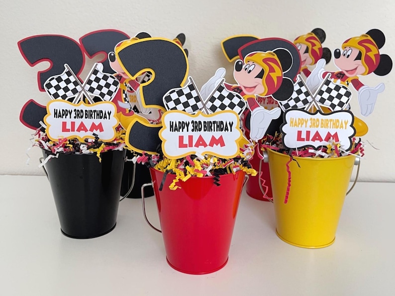 May include: Three Mickey Mouse themed centerpieces for a birthday party. Each centerpiece features a Mickey Mouse character wearing a racing suit, a number three, checkered flags, and a banner that says "Happy 3rd Birthday Liam". The centerpieces are displayed in black, red, and yellow buckets.