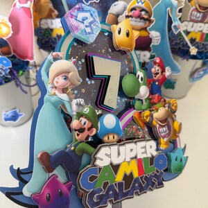 May include: A multi-layered cake topper featuring Super Mario characters including Rosalina, Luigi, Yoshi, and Bowser. The design includes a large number 7, a blue question block, and the text Super Camilo Galaxy in a space-themed font and colours.