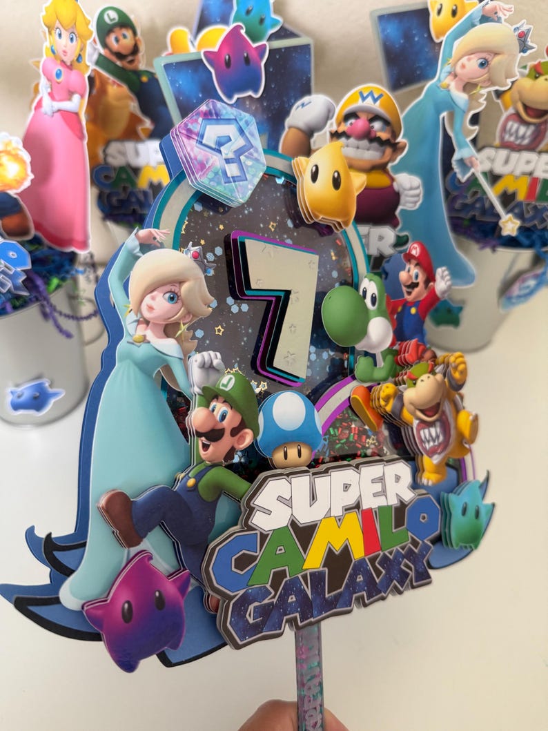 May include: A multi-layered cake topper featuring Super Mario characters including Rosalina, Luigi, Yoshi, and Bowser. The design includes a large number 7, a blue question block, and the text Super Camilo Galaxy in a space-themed font and colours.