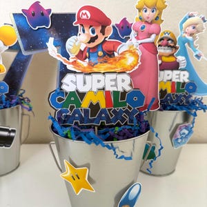 May include: A silver metal bucket centrepiece featuring a cutout of Mario and Princess Peach with the text Super Camilo Galaxy. The bucket is filled with blue and purple paper shreds and decorated with small star and mushroom character stickers.